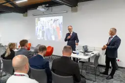 Prezentacja na konferencji z mówcami i uczestnikami w tle.