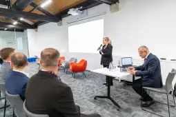 Prezentacja w sali konferencyjnej z osobą mówiącą i słuchaczami.
