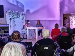 Dwie osoby przy stole podczas występu artystycznego, publiczność w tle.