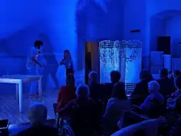 Występ artystyczny w niebieskim świetle, publiczność w tle, interakcja między dwiema osobami.