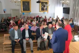 Spotkanie z seniorami w sali, w tle obrazy, uczestnicy w różnych grupach wiekowych.
