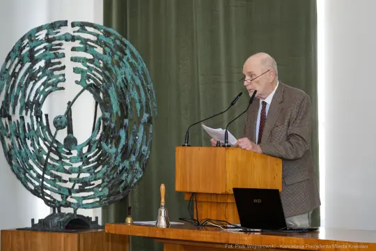Mężczyzna przemawia przy mównicy obok rzeźby w kształcie spirali.