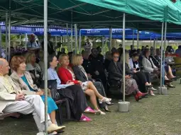 Grupa osób siedzących pod namiotami, uczestniczących w wydarzeniu na świeżym powietrzu.