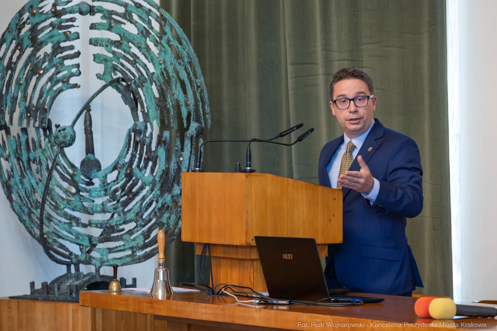 Mężczyzna w garniturze przemawia przy mównicy, obok laptop i dzwonek. W tle abstrakcyjna rzeźba.  Autor: Piotr Wojnarowski