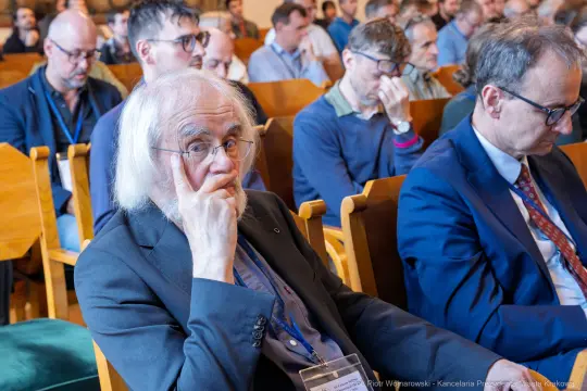 Mężczyzna z siwymi włosami siedzący wśród publiczności na konferencji.