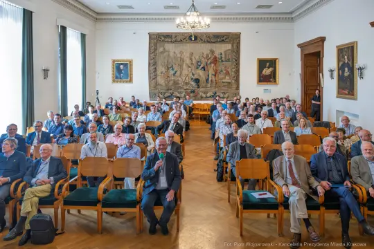 Zgromadzenie ludzi w sali, siedzących na krzesłach, z dekoracyjnym gobelinem w tle.