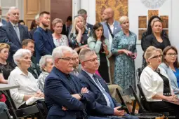 Grupa ludzi w eleganckich strojach, słuchających wystąpienia w sali.