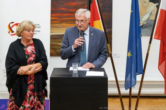 Mężczyzna przemawia przy mównicy, obok stoi kobieta. W tle flagi i obrazy.