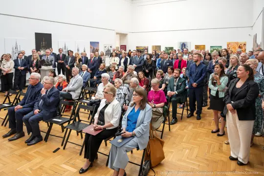 Tłum ludzi w sali wystawowej, siedzących na krzesłach, w eleganckich strojach.