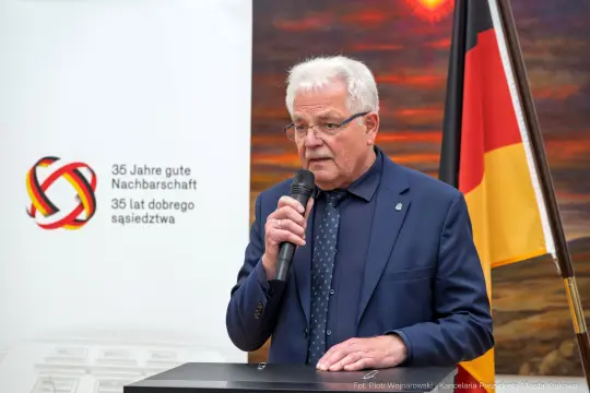 Mężczyzna przemawia przy mównicy, w tle flagi i logo związane z jubileuszem.