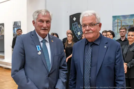 Dwaj mężczyźni w garniturach, jeden z odznaczeniem, podczas wydarzenia kulturalnego.