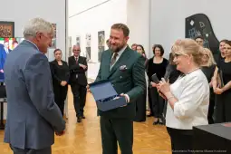 Wręczenie nagrody w galerii sztuki, uśmiechnięci goście w tle.