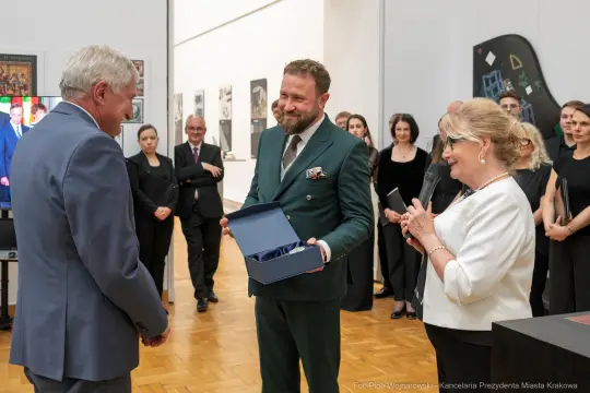Wręczenie nagrody w galerii sztuki, uśmiechnięci goście w tle.
