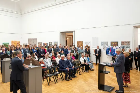 Uroczystość w sali wystawowej z liczną publicznością i przemawiającymi osobami.