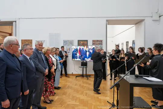 Uroczystość z wystąpieniem i chórem w tle, uczestnicy w eleganckich strojach.