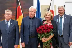 Cztery osoby w eleganckich strojach, jedna z bukietem czerwonych róż, w tle flagi i obrazy.