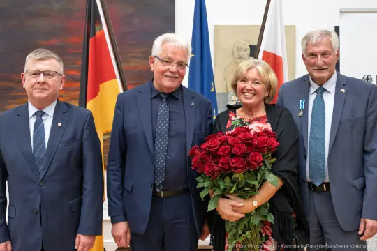 Cztery osoby w eleganckich strojach, jedna z bukietem czerwonych róż, w tle flagi i obrazy.