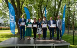 Uczestnicy ceremonii wręczenia nagród stoją na scenie w parku, trzymając dyplomy.