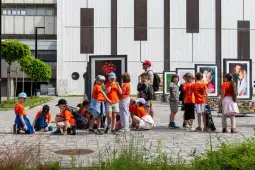 Grupa dzieci w pomarańczowych koszulkach na tle wystawy zdjęć.
