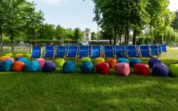 Kolorowe pufy i leżaki w parku otoczone zielenią drzew.