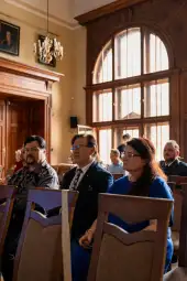 Grupa ludzi siedzących w sali, z dużymi oknami i eleganckim wystrojem.