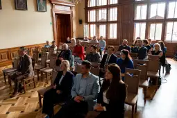 Grupa ludzi siedząca w sali z drewnianymi ścianami i dużymi oknami.