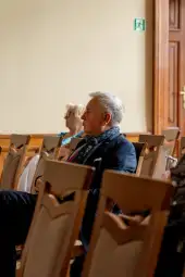 Mężczyzna w garniturze siedzący na krześle w sali, w tle kobieta z jasnymi włosami.