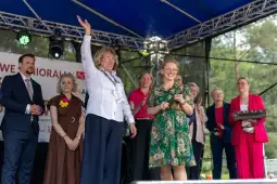 Kobieta z uniesioną ręką na scenie, obok grupy uśmiechniętych osób.