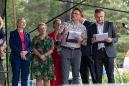 Grupa ludzi na scenie, w różnych strojach, trzymających dokumenty i mikrofon.