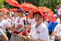 Grupa kobiet w białych koszulkach z czerwonymi parasolkami, uśmiechających się i trzymających telefon.