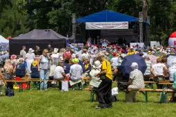 Tłum ludzi w białych koszulkach na wydarzeniu plenerowym z występami na scenie.