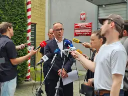 Polityk udziela wywiadu dziennikarzom przed budynkiem z flagą Polski.