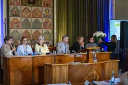 Panel dyskusyjny z sześcioma osobami siedzącymi przy stole, jedna osoba wręcza bukiet kwiatów.