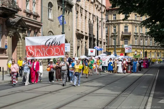 Parada seniorów w kolorowych strojach na ulicach Krakowa. Transparenty i balony w tle.