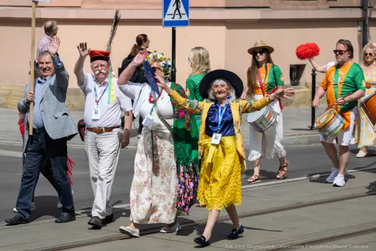 Grupa osób w kolorowych strojach maszeruje ulicą, machając rękami i uśmiechając się.
