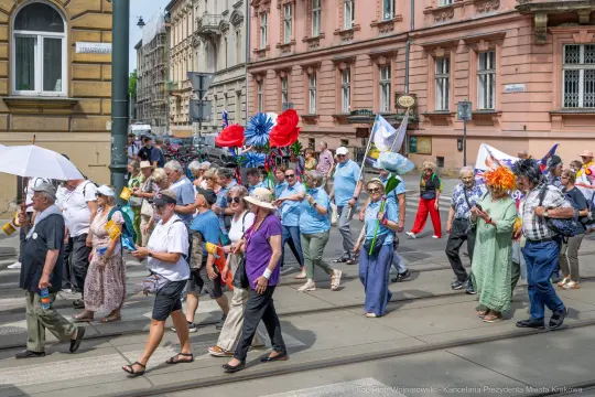 Tłum ludzi w kolorowych strojach, niosących flagi i parasole, idzie ulicą podczas parady.