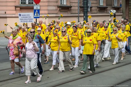 Grupa osób w żółtych koszulkach z kwiatami na głowach maszeruje ulicą.