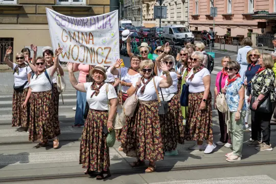 Grupa kobiet w tradycyjnych strojach, trzymających transparent, pozdrawia przechodniów.