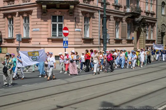 Grupa ludzi idąca w marszu, trzymająca flagi i transparenty, w tle architektura miasta.