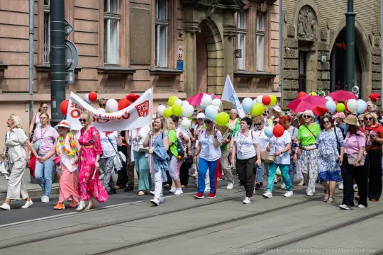 Grupa ludzi z balonami i transparentami uczestniczy w marszu ulicami miasta.