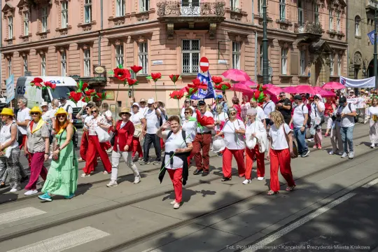 Grupa ludzi w białych i czerwonych strojach, niosących różowe parasole i kwiaty, maszeruje ulicą.