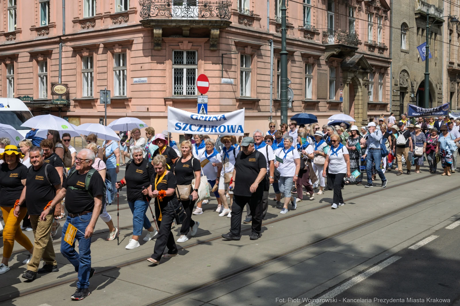 Grupa ludzi w koszulkach z logo, maszerująca w słońcu z parasolami, niosąca transparent.  Autor: Piotr Wojnarowski