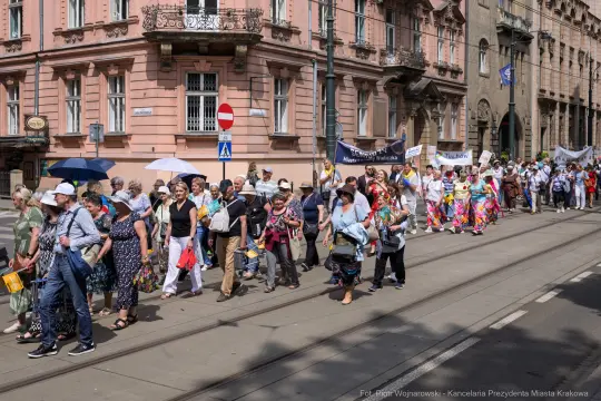 Grupa ludzi w kolorowych strojach maszeruje ulicą, niosąc transparenty i parasole.