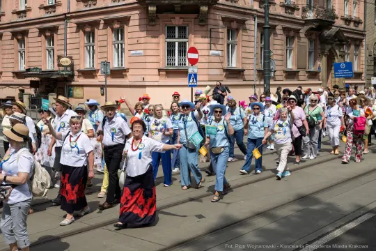 Grupa ludzi w kolorowych strojach maszeruje ulicą, niektórzy machają rękami.