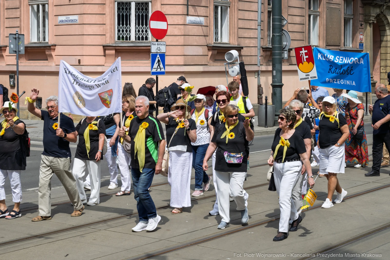 Seniorzy w białych strojach z żółtymi kwiatami, maszerujący w parku, trzymający transparenty.  Autor: Piotr Wojnarowski