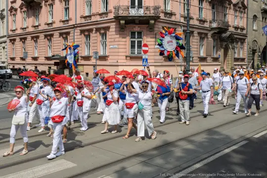 Grupa ludzi w białych strojach z parasolkami i kolorowymi akcesoriami podczas parady.