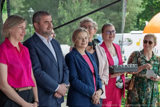Grupa osób na wydarzeniu, uśmiechnięte, w eleganckich strojach, z różnymi akcesoriami.