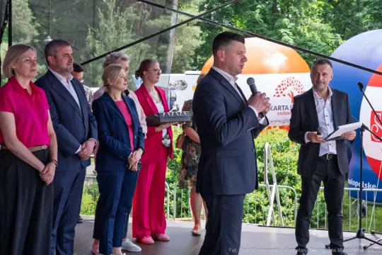 Mężczyzna przemawia na scenie, obok grupy osób w formalnych strojach.