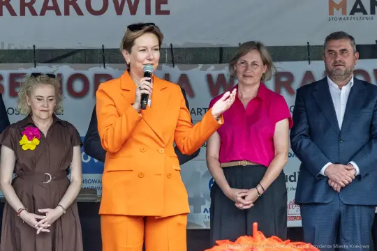 Kobieta w pomarańczowym garniturze przemawia do zgromadzonego tłumu, obok niej stoją inne osoby.