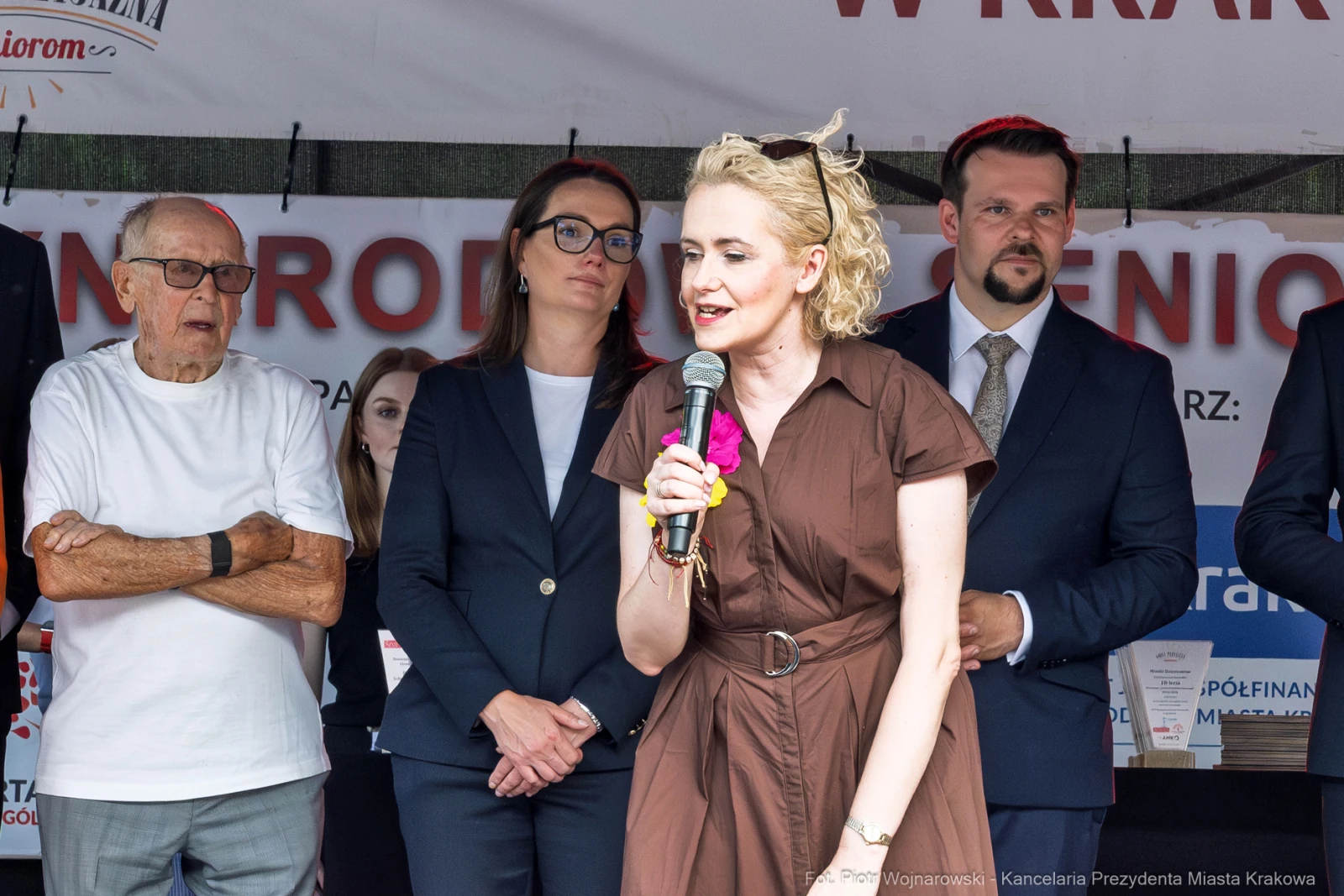 Kobieta w brązowej sukience przemawia na tle grupy ludzi podczas wydarzenia publicznego.  Autor: Piotr Wojnarowski
